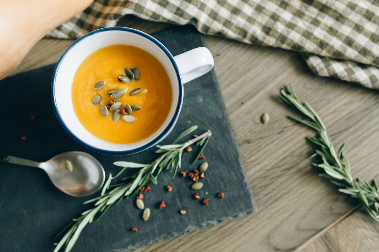 Top view of creamy pumpkin soup in a cup with rosemary and seeds on a slate.